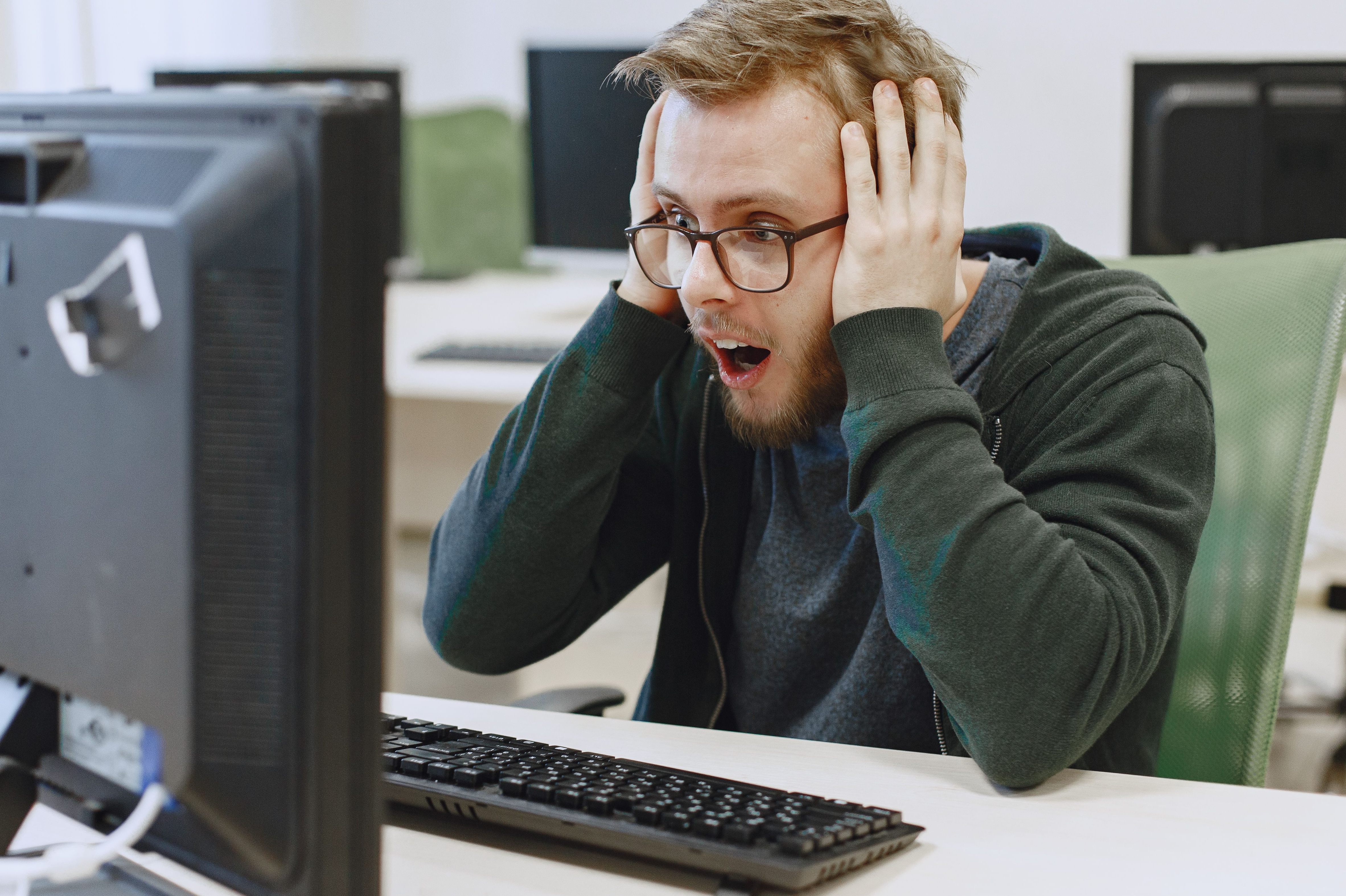 Man wearing glasses stares at a computer monitor with his mouth open and both hands holding the sides of his head.
