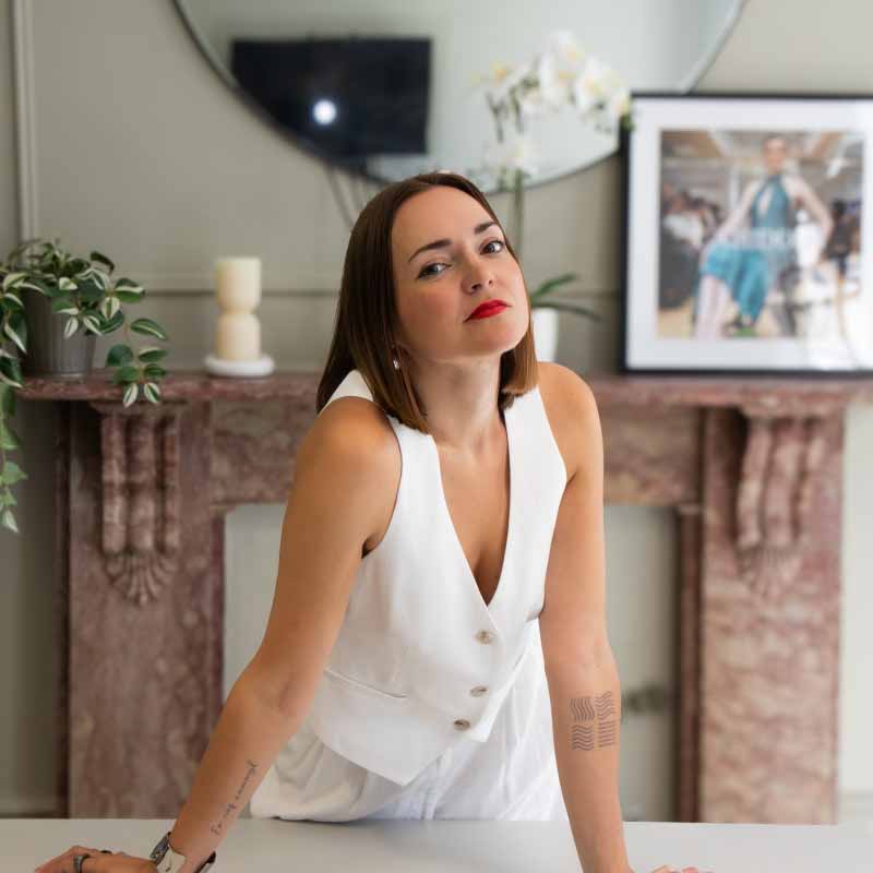 Victoria Jenkins wearing an armless white woman's suit standing in front of a fireplace
